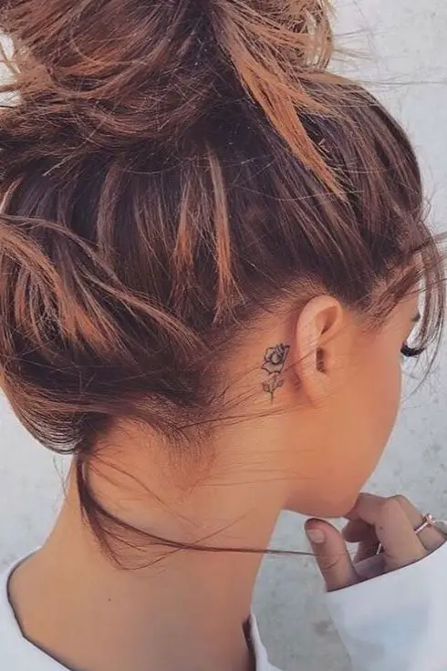 Chica con el cabello recogido mostrando el tatuaje de una rosa pequeña detrás de su oreja